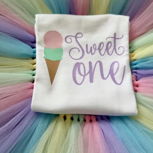 May include: A white t-shirt featuring a graphic of an ice cream cone with a pink scoop and a green scoop, accompanied by the purple text "Sweet one" in a cursive font, layered over a multi-colored pastel tulle skirt with pink, yellow, and blue.