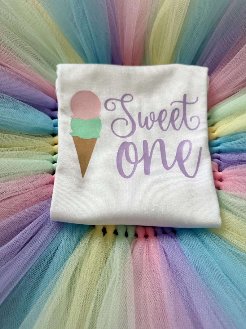 May include: A white t-shirt featuring a graphic of an ice cream cone with a pink scoop and a green scoop, accompanied by the purple text "Sweet one" in a cursive font, layered over a multi-colored pastel tulle skirt with pink, yellow, and blue.
