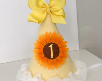 May include: A yellow cone-shaped party hat with a large yellow bow at the top. The hat features a felt sunflower with a brown center and the number 1. A white ruffled trim and a white ribbon complete the design.