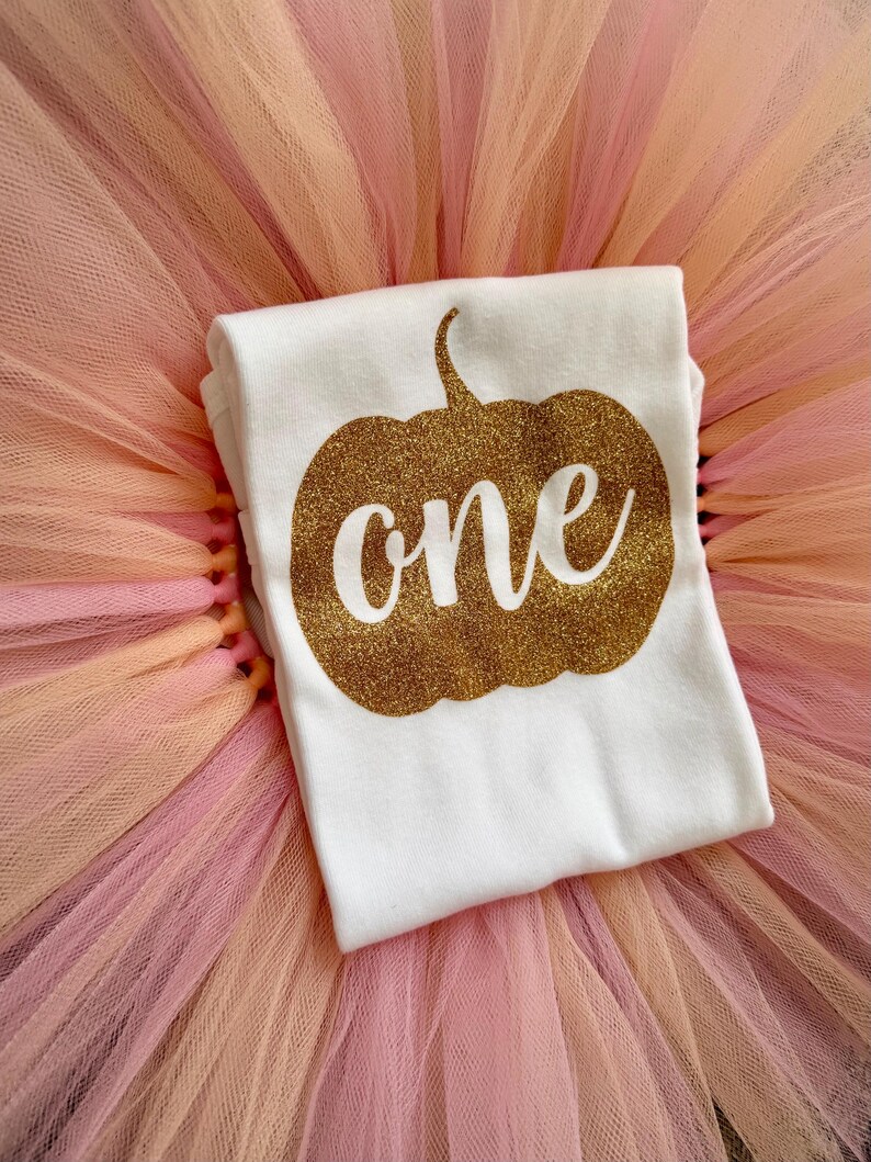 May include: A white t-shirt featuring a gold glitter pumpkin graphic with the word one written in white script across the center, layered over a fluffy, multi-layered tulle skirt in shades of soft peach, light pink, and pale orange.