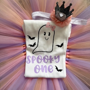 May include: A white t-shirt featuring a black ghost graphic with a pink bow, black bats, and the purple text SPOOKY one. The shirt is paired with a multi-layered tulle tutu in shades of peach and lavender, and a headband with a small crown.