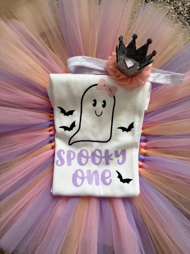 May include: A white t-shirt featuring a black ghost graphic with a pink bow, black bats, and the purple text SPOOKY one. The shirt is paired with a multi-layered tulle tutu in shades of peach and lavender, and a headband with a small crown.