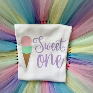 May include: A white t-shirt featuring a graphic of an ice cream cone with a pink scoop and a green scoop, accompanied by the purple text "Sweet one". The shirt is layered over a multi-colored pastel tulle skirt with shades of pink, yellow, and blue.