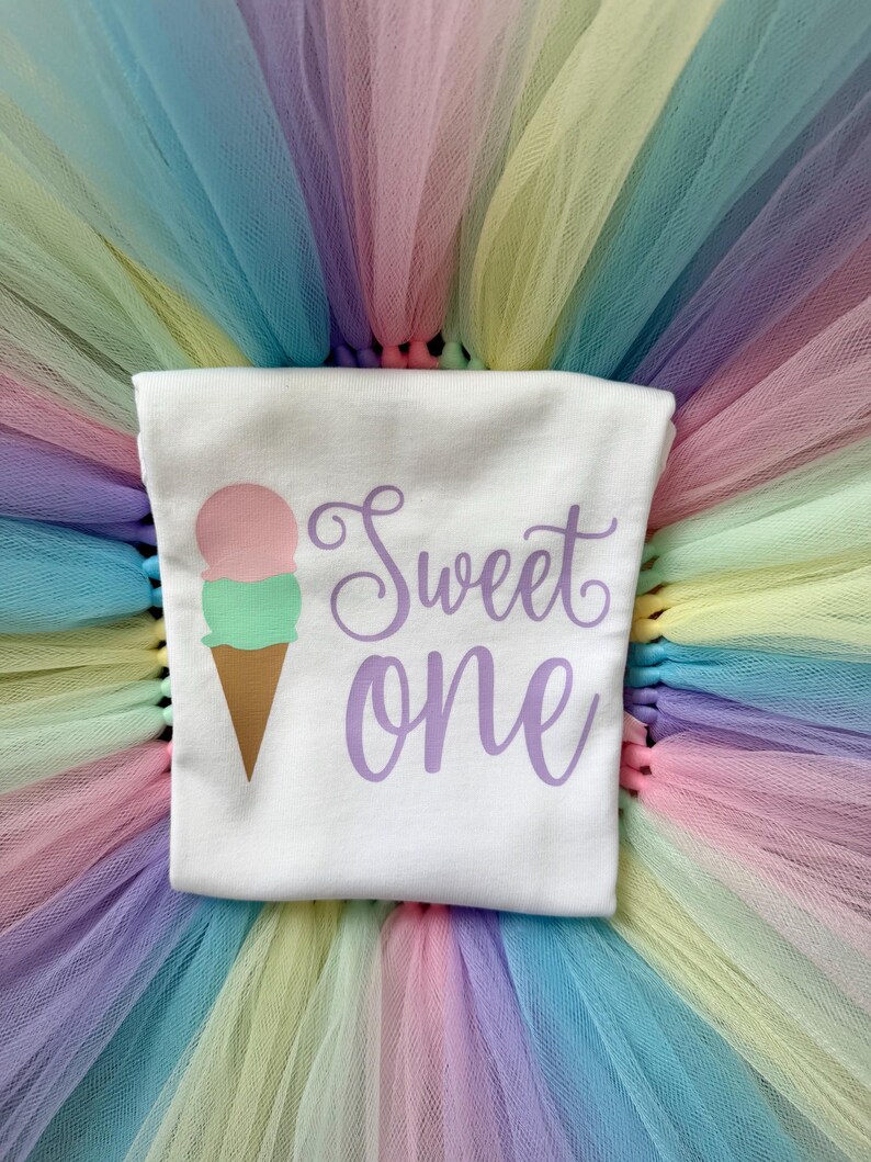 May include: A white t-shirt featuring a graphic of an ice cream cone with a pink scoop and a green scoop, accompanied by the purple text "Sweet one". The shirt is layered over a multi-colored pastel tulle skirt with shades of pink, yellow, and blue.