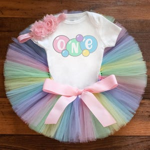 May include: A white short-sleeved bodysuit with the text ONE in colorful bubbles, paired with a multi-colored pastel tulle tutu featuring a large pink satin bow at the waist and a matching pink floral headband with two fabric flowers.