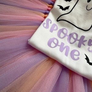 May include: A white t-shirt featuring a black line-art ghost and bat graphic with the purple text SPOOKY one, layered over a multi-layered tulle tutu skirt in alternating pastel shades of lavender, peach, and light pink fabric strips.