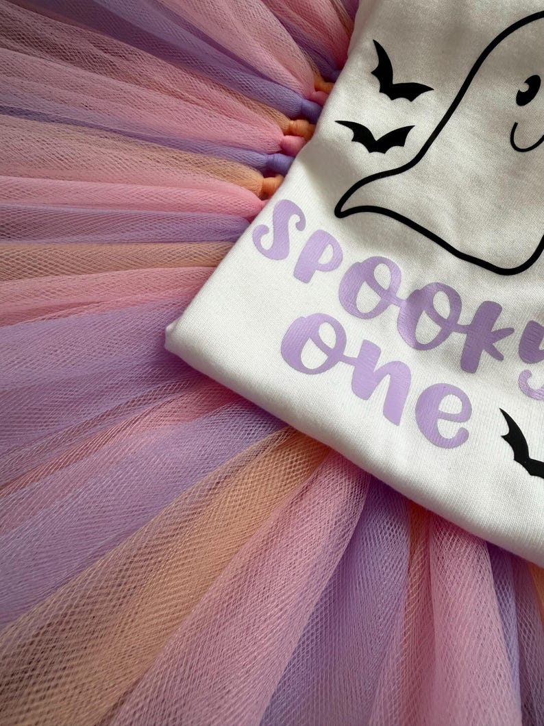 May include: A white t-shirt featuring a black line-art ghost and bat graphic with the purple text SPOOKY one, layered over a multi-layered tulle tutu skirt in alternating pastel shades of lavender, peach, and light pink fabric strips.