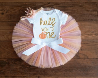 May include: A white onesie with the text "half way to one" and a pumpkin graphic. The onesie is paired with a peach and pink tulle tutu with a white satin bow.