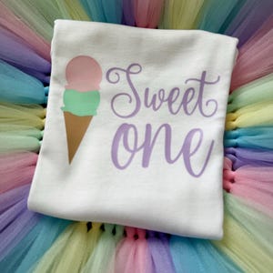 May include: A white t-shirt featuring a graphic of a two-scoop ice cream cone with a pink top scoop and a green bottom scoop. The text "Sweet one" is printed in a purple cursive font to the right of the ice cream cone graphic.
