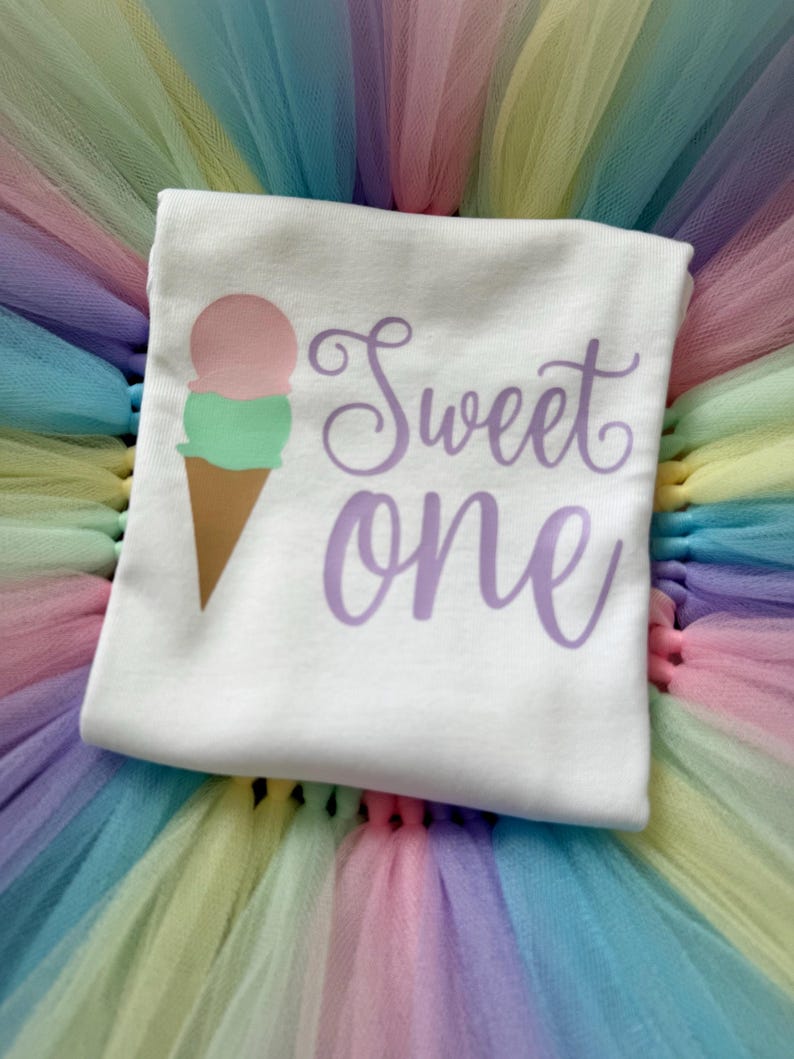 May include: A white t-shirt featuring a graphic of a two-scoop ice cream cone with a pink top scoop and a green bottom scoop. The text "Sweet one" is printed in a purple cursive font to the right of the ice cream cone graphic.