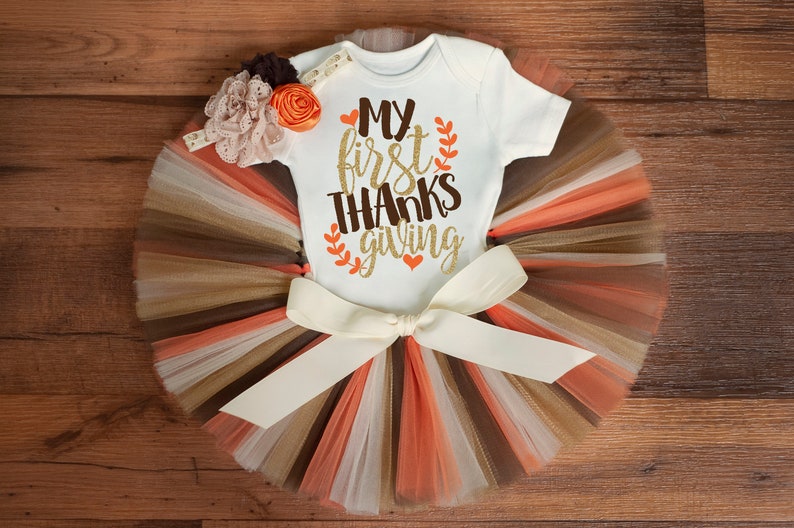 May include: A white baby onesie with the text "My first THANKSgiving" in gold glitter lettering and orange leaves. The onesie is paired with a brown, beige, and orange tulle tutu with a white satin bow.