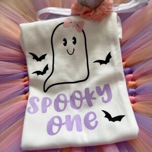 May include: A white t-shirt featuring a black outline of a ghost with a pink bow, three black bats, and the purple text "SPOOKY one". The shirt is paired with a glittery crown headband adorned with a peach-coloured fabric flower and a multi-coloured tulle skirt.