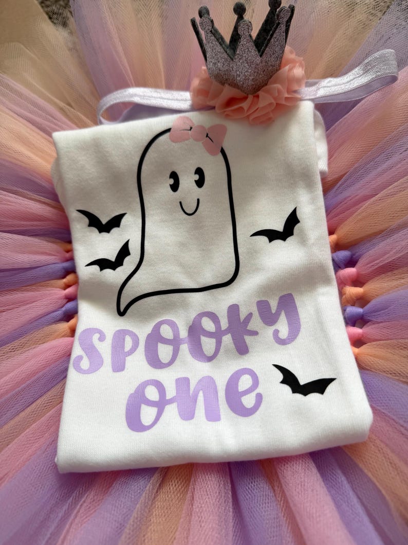 May include: A white t-shirt featuring a black outline of a ghost with a pink bow, three black bats, and the purple text "SPOOKY one". The shirt is paired with a glittery crown headband adorned with a peach-coloured fabric flower and a multi-coloured tulle skirt.