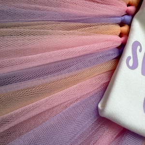 May include: A layered tulle skirt featuring alternating strips of pastel purple, soft pink, and peach fabric. Attached to the side is a white fabric panel with a portion of purple text visible, showing the letter S and part of another character.