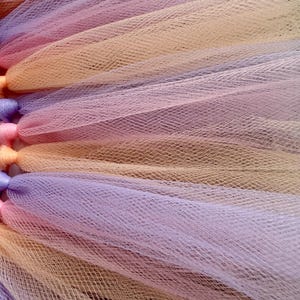 May include: A layered tulle skirt featuring alternating strips of soft peach, lavender, and light pink fabric. The material has a fine, mesh-like texture with gathered sections at the top, creating a voluminous and pleated appearance throughout.