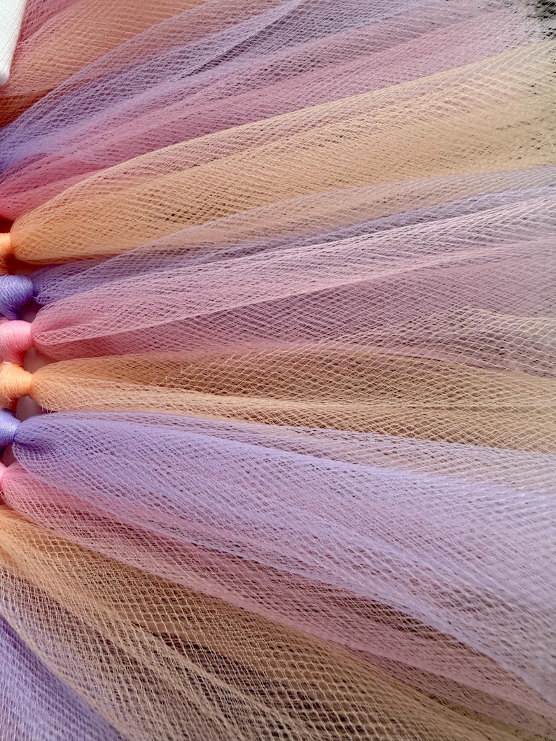 May include: A layered tulle skirt featuring alternating strips of soft peach, lavender, and light pink fabric. The material has a fine, mesh-like texture with gathered sections at the top, creating a voluminous and pleated appearance throughout.