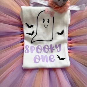 May include: A white t-shirt featuring a black ghost graphic with a pink bow, black bats, and the purple text "SPOOKY one". The shirt is paired with a pastel multi-coloured tulle tutu and a headband with a peach-coloured flower and a silver crown.