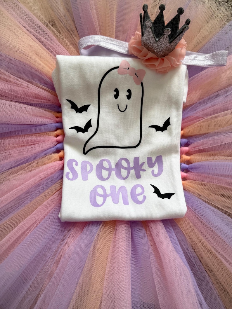 May include: A white t-shirt featuring a black ghost graphic with a pink bow, black bats, and the purple text "SPOOKY one". The shirt is paired with a pastel multi-coloured tulle tutu and a headband with a peach-coloured flower and a silver crown.