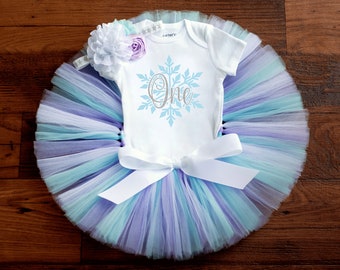 May include: A white onesie with a blue snowflake and the word "One" in silver glitter. The onesie is paired with a light blue, purple, and white tulle tutu with a white satin bow. A white headband with a white flower and a purple flower is included.