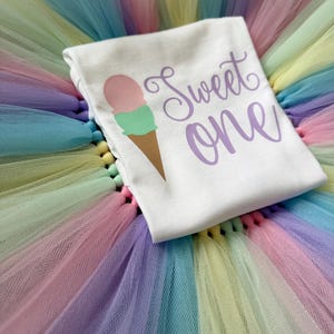 May include: A white t-shirt featuring a graphic of an ice cream cone with a pink scoop and a green scoop, accompanied by the purple text "Sweet one" in a cursive font, resting on a layered tulle skirt with pastel pink, blue, yellow, and purple colors.