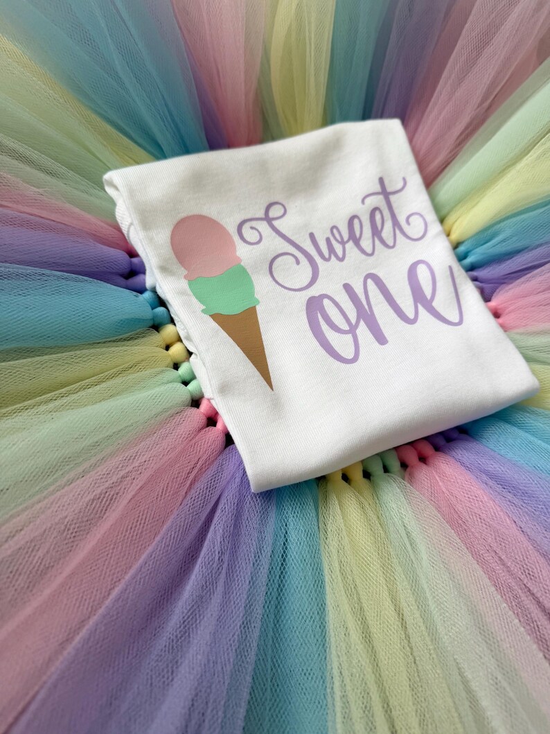 May include: A white t-shirt featuring a graphic of an ice cream cone with a pink scoop and a green scoop, accompanied by the purple text "Sweet one" in a cursive font, resting on a layered tulle skirt with pastel pink, blue, yellow, and purple colors.