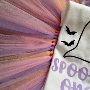 May include: A white short-sleeved bodysuit featuring a black ghost graphic with two small black bats and the purple text "SPOOKY one" paired with a layered tulle tutu skirt in alternating shades of pastel pink, lavender, and peach.