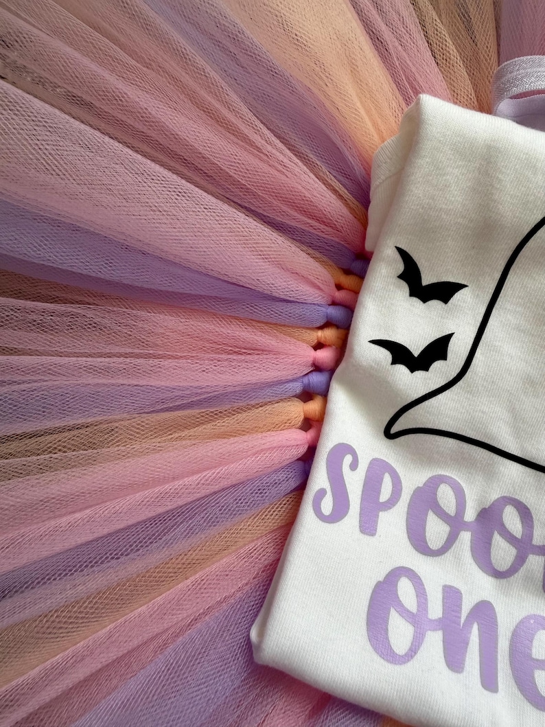 May include: A white short-sleeved bodysuit featuring a black ghost graphic with two small black bats and the purple text "SPOOKY one" paired with a layered tulle tutu skirt in alternating shades of pastel pink, lavender, and peach.