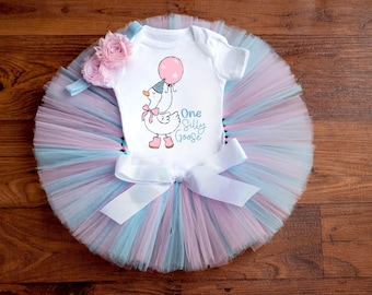 May include: A white onesie with a pink balloon and a goose with a party hat, the text "One Silly Goose" is printed on the onesie. The onesie is paired with a pink and blue tulle tutu with a white satin bow.