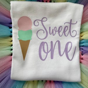 May include: A white t-shirt featuring a graphic of an ice cream cone with a pink scoop and a green scoop. To the right of the ice cream cone, the text "Sweet one" is printed in a purple cursive font. The shirt is placed on a pastel-colored tulle fabric.