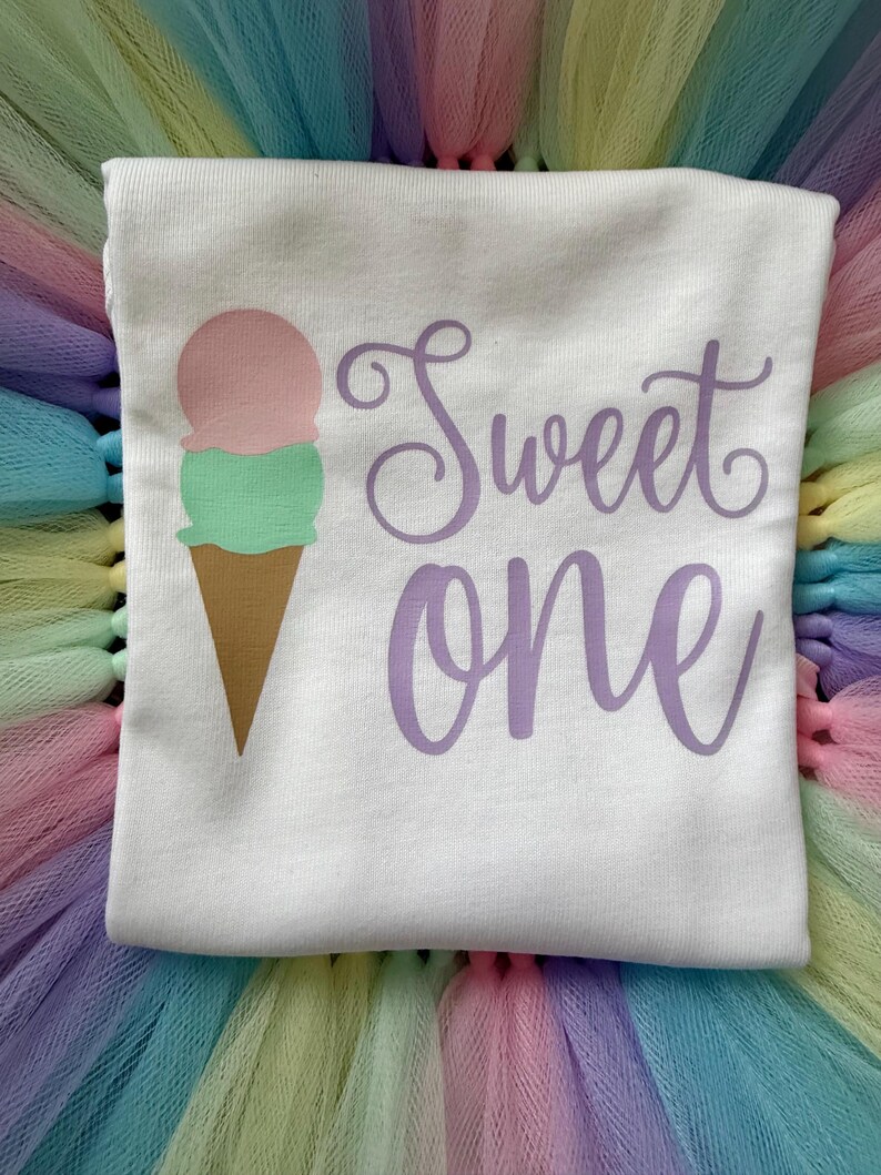 May include: A white t-shirt featuring a graphic of an ice cream cone with a pink scoop and a green scoop. To the right of the ice cream cone, the text "Sweet one" is printed in a purple cursive font. The shirt is placed on a pastel-colored tulle fabric.