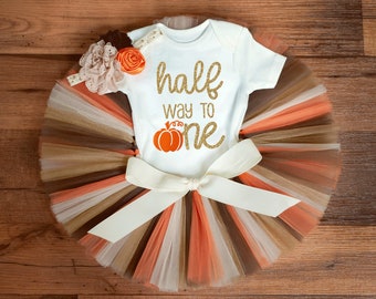 May include: A white onesie with gold glitter text that reads "half way to one" and a small orange pumpkin graphic. The onesie is paired with a brown, beige, and orange tulle tutu with a white satin bow.