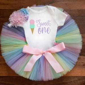 May include: A white short-sleeved bodysuit with the text Sweet one and an ice cream cone graphic, paired with a multi-colored pastel tulle tutu featuring a large pink satin bow at the waist and a matching floral headband with pink and blue petals.