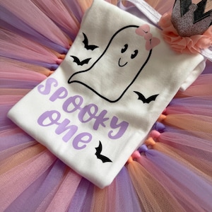 May include: A white t-shirt featuring a black outline of a ghost with a pink bow, three black bats, and the purple text "Spooky one". The shirt is placed on a multi-coloured tulle skirt and includes a small grey crown accessory with a peach ruffle.