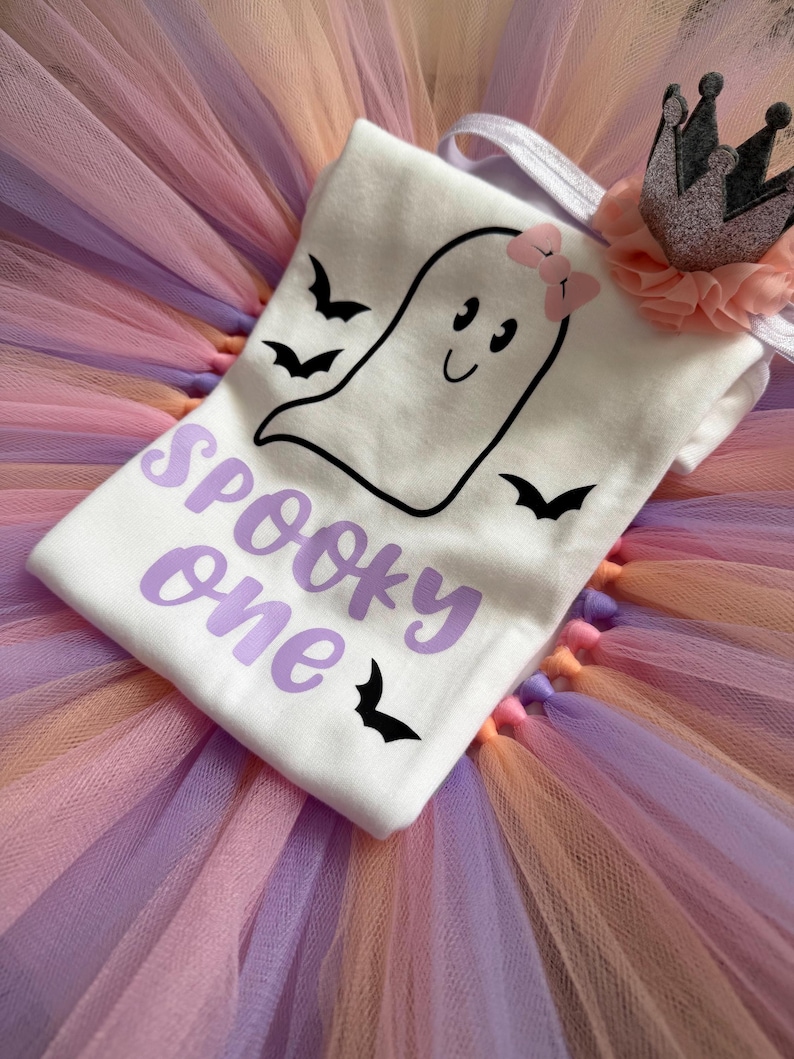 May include: A white t-shirt featuring a black outline of a ghost with a pink bow, three black bats, and the purple text "Spooky one". The shirt is placed on a multi-coloured tulle skirt and includes a small grey crown accessory with a peach ruffle.