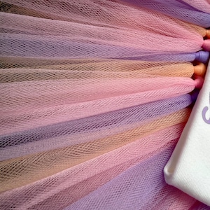 May include: A layered tulle skirt featuring alternating strips of light purple, soft peach, and pale pink mesh fabric. The tulle is gathered and attached to a white fabric waistband that displays a portion of purple text on its surface.