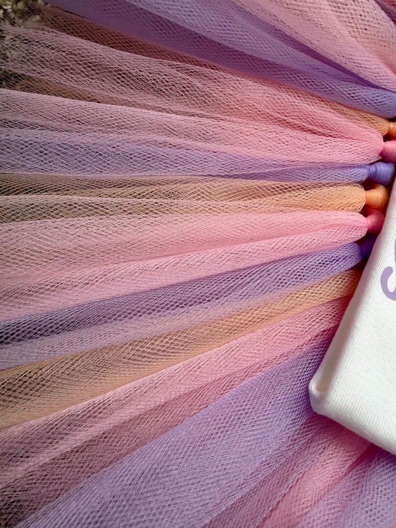 May include: A layered tulle skirt featuring alternating strips of light purple, soft peach, and pale pink mesh fabric. The tulle is gathered and attached to a white fabric waistband that displays a portion of purple text on its surface.