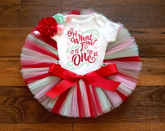 May include: A white short-sleeved bodysuit with the red and green text Oh What Fun It Is To Be One, paired with a multi-layered tulle tutu in red, white, and mint green, featuring a large red satin bow and a cluster of red and mint fabric flowers.