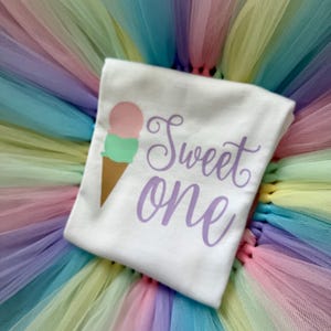May include: A white t-shirt featuring a graphic of an ice cream cone with a pink scoop and a green scoop, accompanied by the purple script text "Sweet one". The shirt is placed on a multi-colored pastel tulle skirt with layers of pink, yellow, and blue.