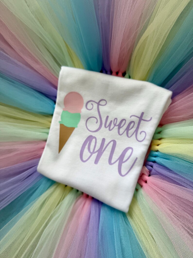 May include: A white t-shirt featuring a graphic of an ice cream cone with a pink scoop and a green scoop, accompanied by the purple script text "Sweet one". The shirt is placed on a multi-colored pastel tulle skirt with layers of pink, yellow, and blue.