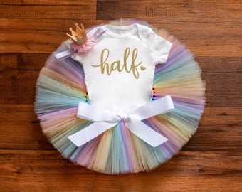 May include: A white baby bodysuit with the word "half" in gold script, set on a pastel rainbow tutu. A small gold crown and a white bow complete the outfit, ideal for a first birthday celebration.