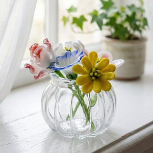 May include: A clear glass vase with a ribbed design contains colourful glass flowers. The flowers feature red, white, blue, and yellow petals. The vase is on a white windowsill, with a sheer curtain and a potted plant in the background.