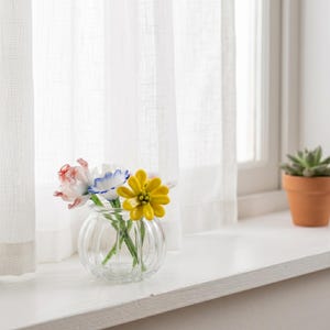 May include: A clear glass vase with a ribbed design contains a colourful arrangement of glass flowers. The flowers include a pink and white tulip, a blue and white flower, and a yellow daisy. The vase sits on a white windowsill near a window with sheer white curtains.