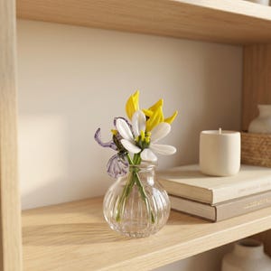 May include: A small, clear glass vase with a ribbed design contains an arrangement of artificial flowers. The flowers have white, yellow, and purple petals. The vase is on a wooden shelf with books and a candle.