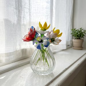 May include: A clear glass vase holding a bouquet of glass flowers in various colours. The flowers include red, blue, yellow, and white petals. The vase is on a white windowsill, with a small potted plant in the background.