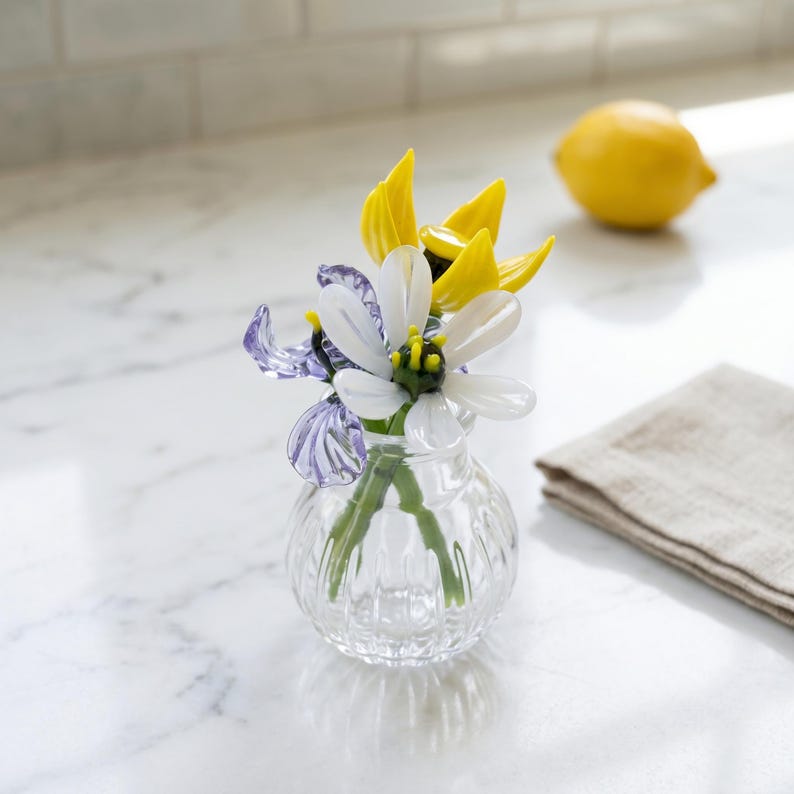 May include: A small, clear glass vase with a ribbed design holds a colourful arrangement of glass flowers. The flowers include white, yellow, and purple petals with green and yellow centres. A yellow lemon and a beige linen napkin are in the background.