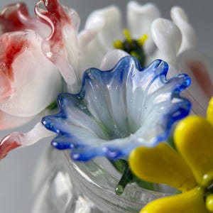 May include: A close-up of a glass flower arrangement in a clear jar. The flowers are various colours, including white, red, blue, and yellow. The blue flower is in focus, with a ruffled edge. The arrangement is set against a soft, neutral background.