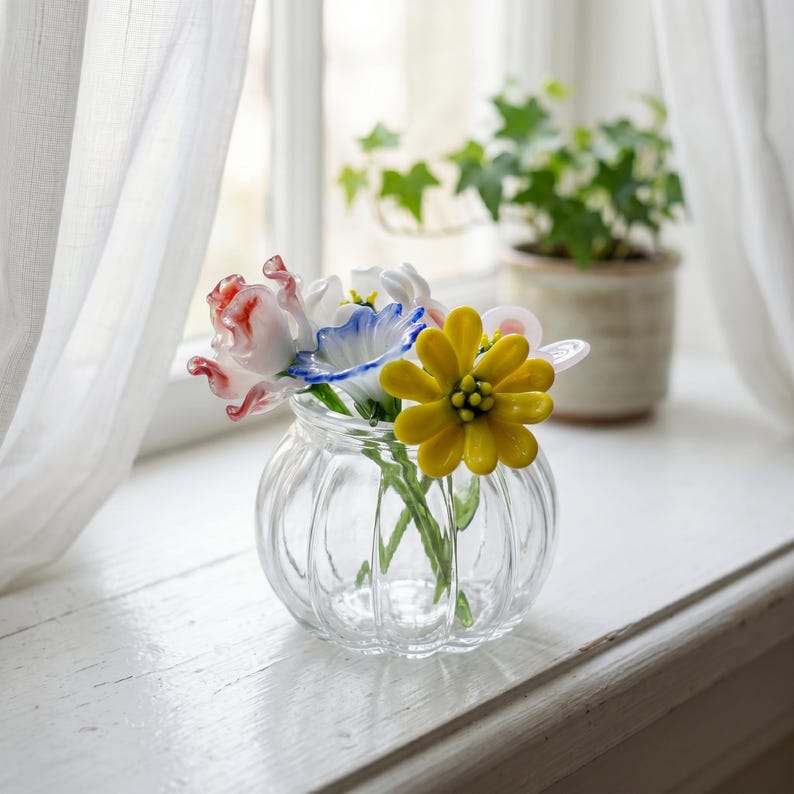 May include: A clear glass vase with a ribbed design contains colourful glass flowers. The flowers feature red, white, blue, and yellow petals. The vase is on a white windowsill, with a sheer curtain and a potted plant in the background.