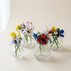 May include: Five small, clear glass vases, each containing a different arrangement of colourful, artificial flowers. The flowers include yellow, white, red, and blue blooms. The vases have a ribbed design and are arranged on a neutral background.