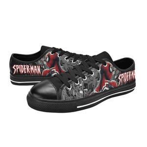 May include: Black canvas sneakers featuring a Spider-Man design. The shoes have a black toe cap, black laces, and a white stripe along the sole. The Spider-Man graphic is red and black, set against a gray cityscape.