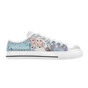 May include: A low-top canvas sneaker featuring a light blue graphic print of characters from the movie Frozen, including Elsa and Anna, with the text FROZEN visible on the side. The shoe has white laces, a white rubber toe cap, and a white sole.